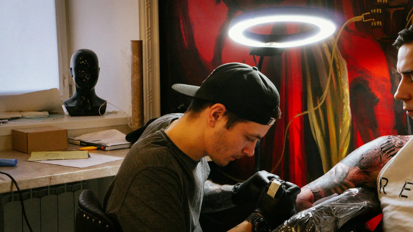 A tattoo artist concentrating on a client’s arm beside a window.